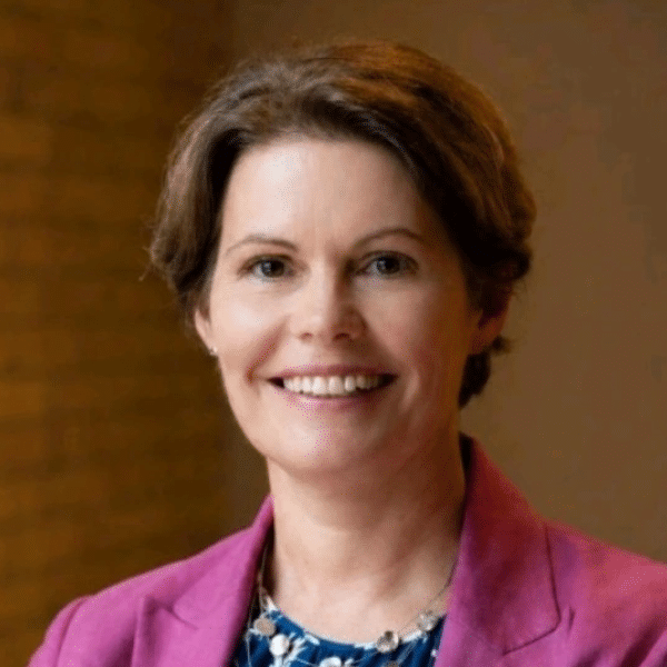 Smiling woman in a pink blazer and blue floral blouse, standing against a brick wall backdrop and looking at the camera.