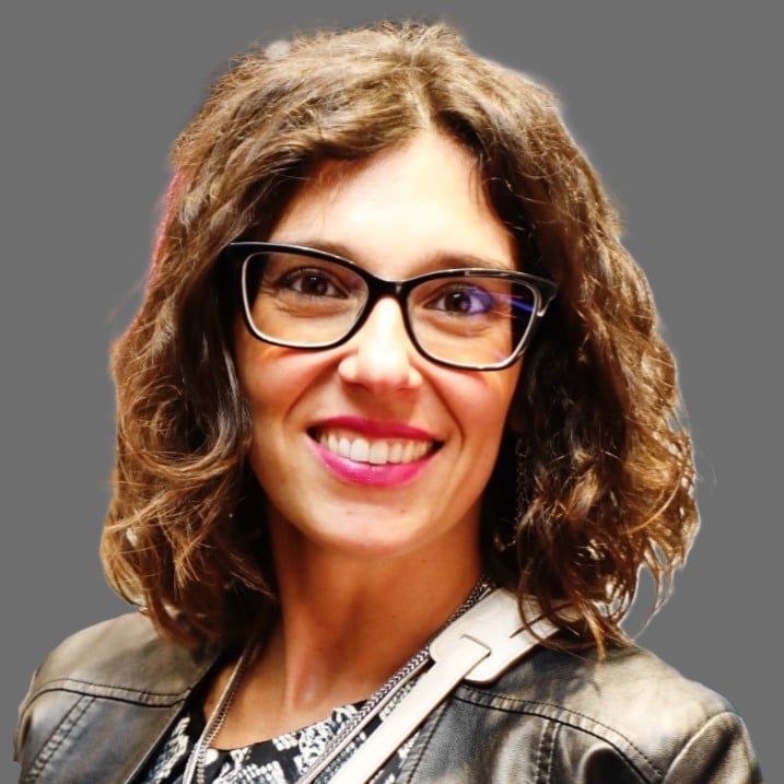 Smiling woman with curly brown hair and black-framed glasses, wearing a leather jacket against a gray background.