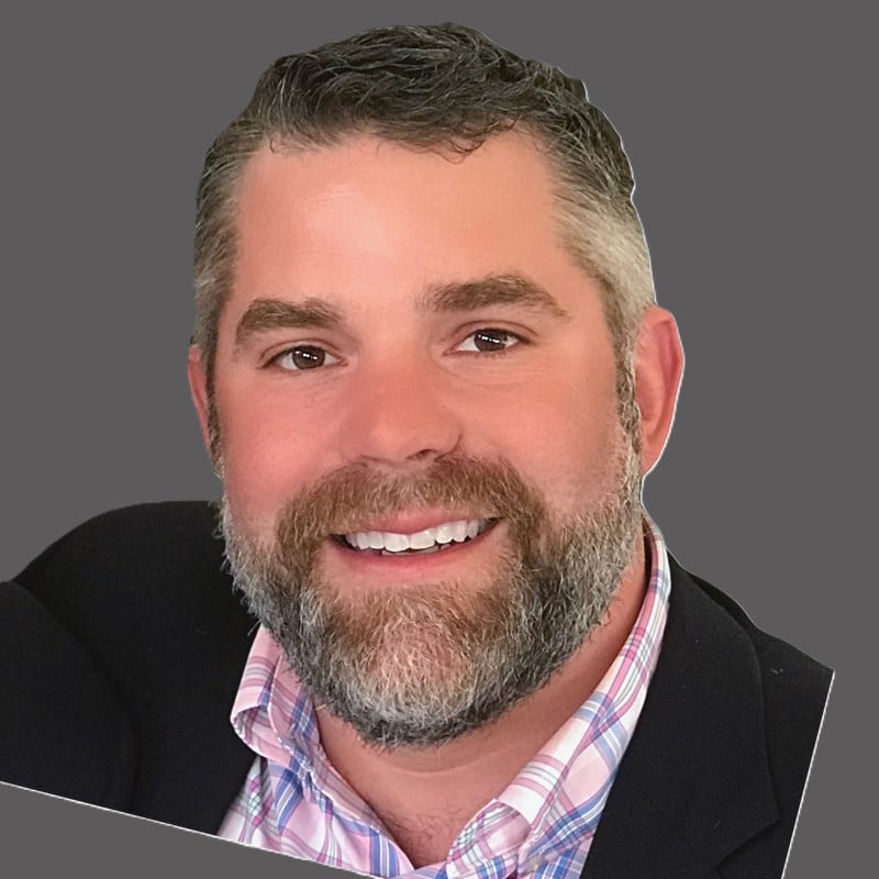 Smiling man with a full beard and short gray-streaked hair in a pink plaid shirt and dark blazer against a gray background